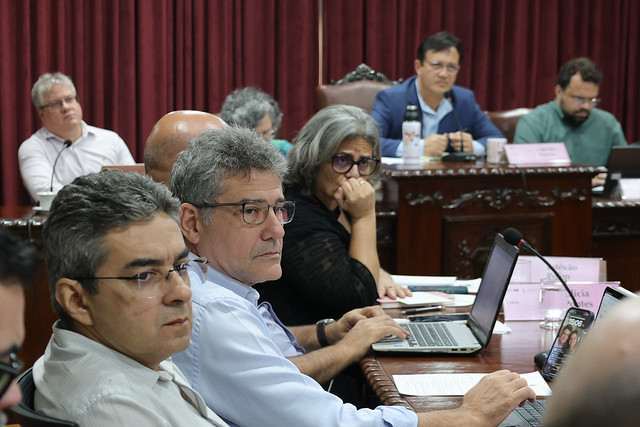 Imagem: pessoas sentadas ao redor de uma grande mesa de madeira, em ambiente de reunião formal. Em primeiro plano, três homens olham atentos para frente, dois deles usando óculos e com notebooks abertos. Mais ao fundo, uma mulher de cabelos grisalhos e óculos apoia a mão no rosto, também concentrada. Outras pessoas aparecem na mesa, algumas com computadores e papéis, todas voltadas para a discussão. Ao fundo, cortinas vermelhas escuras cobrem a parede.