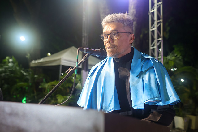 Imagem: homem de cabelos grisalhos e óculos redondos fala ao microfone durante cerimônia noturna. Ele veste beca preta com capa azul clara sobre os ombros. Ao fundo, há árvores iluminadas e parte de uma tenda branca, compondo o cenário do evento ao ar livre.