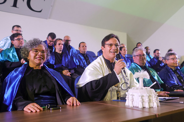 Imagem: mesa de honra de uma cerimônia de colação de grau, com vários docentes vestidos de beca preta e capas azul-escuras. Em destaque, uma mulher de óculos e cabelos grisalhos cacheados sorri, enquanto um homem ao seu lado, também de óculos, usa capa branca e fala ao microfone. À frente deles, sobre a mesa, há uma escultura branca em formato de jangada com vela. Ao fundo, outros professores acompanham a solenidade