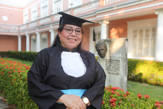 Imagem: mulher branca, de cabelos pretos e lisos, posa sorrindo para foto, vestindo beca, capelo, faixa azul e usando óculos. Ao fundo, aparece a fachada cor de rosa do prédio da Reitoria da UFC