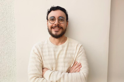 Imagem: homem branco vestindo camisa listrada, de braços cruzados e encostado na parede