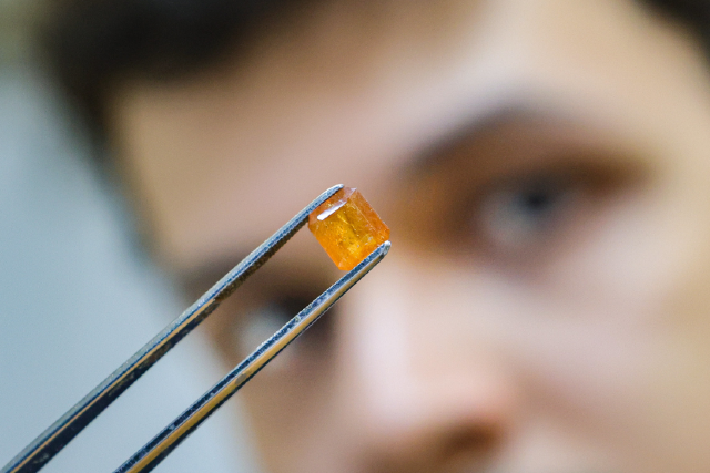 Imagem: Fotografia em close-up de uma pessoa segurando uma pequena pinça metálica. A pinça segura um cristal laranja e translúcido, em formato cúbico. O rosto da pessoa aparece desfocado ao fundo, destacando o mineral como principal foco da imagem.
