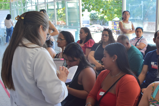 Imagem: Em um espaço amplo e iluminado, uma profissional de jaleco branco segura um caderno e conversa com um grupo de pessoas sentadas. As pessoas, em sua maioria mulheres, prestam atenção à profissional enquanto aguardam atendimento. Ao fundo, há mais pessoas em pé próximas às janelas de vidro, por onde entra a luz natural e se vê parte de árvores e construções externas.