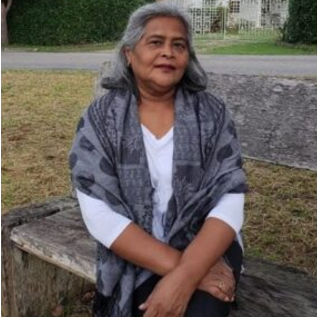 Imagem: mulher de pele morena e cabelos grisalhos, soltos e lisos, está sentada em um banco de madeira rústico ao ar livre. Ela veste blusa branca de manga 3/4 e está coberta com um xale cinza estampado em tons escuros. A mulher cruza as pernas e mantém as mãos sobre o colo, olhando levemente para a frente com expressão serena. Ao fundo, há uma rua pavimentada, grama seca e uma cerca viva verde