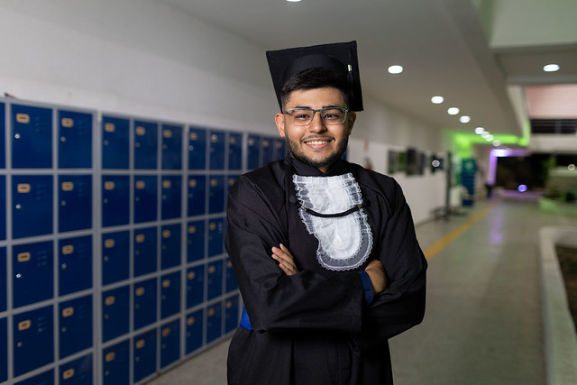 Imagem: Homem jovem, usando óculos, beca e capelo pretos, sorri para a câmera com os braços cruzados. Ele está em um corredor iluminado, ao lado de armários azuis alinhados na parede. A cena transmite clima de celebração de formatura.