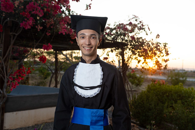 Imagem: Homem jovem veste beca preta com faixa azul e capelo de formatura. Ele sorri para a câmera em um jardim, com flores cor-de-rosa e o sol se pondo ao fundo, criando um clima de celebração e conquista.