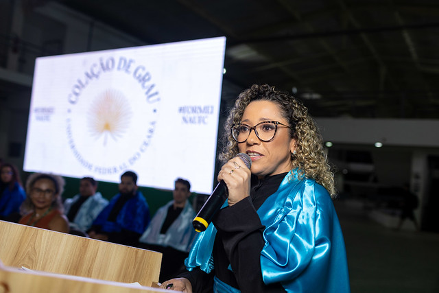 Imagem: Mulher parda, de cabelos encaracolados, usando óculos e beca azul brilhante fala ao microfone durante uma cerimônia de colação de grau da Universidade Federal do Ceará. Ela está em pé atrás de um púlpito de madeira, e ao fundo aparece um telão com o logotipo da universidade e as palavras “Colação de Grau”. Algumas pessoas de beca estão sentadas no palco.
