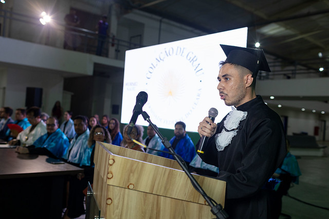 Imagem: Homem jovem vestido com beca e capelo pretos fala ao microfone em um púlpito de madeira durante cerimônia de colação de grau da Universidade Federal do Ceará. Atrás dele, no palco, estão várias pessoas usando becas azuis, e ao fundo aparece um telão iluminado com o logotipo da universidade e o texto “Colação de Grau”