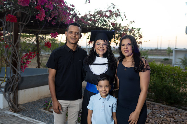 Imagem: Mulher parda vestida com beca e capelo de formatura sorri ao lado de três familiares: um homem, uma mulher e uma criança. Eles posam juntos em um jardim com flores e árvores ao fundo, celebrando a conquista acadêmica.