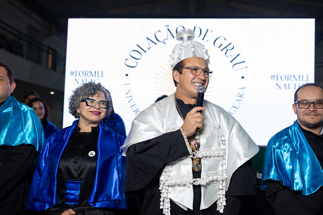 Imagem: Cerimônia de colação de grau em uma universidade. No centro da imagem, um homem veste beca preta com faixa e capa brancas ornamentadas, além de um chapéu tradicional acadêmico, segurando um microfone e sorrindo enquanto fala. Ao lado dele, estão autoridades acadêmicas vestindo becas pretas com capas azuis brilhantes. Ao fundo, um painel iluminado exibe as palavras “Colação de Grau – Universidade Federal do Ceará”.