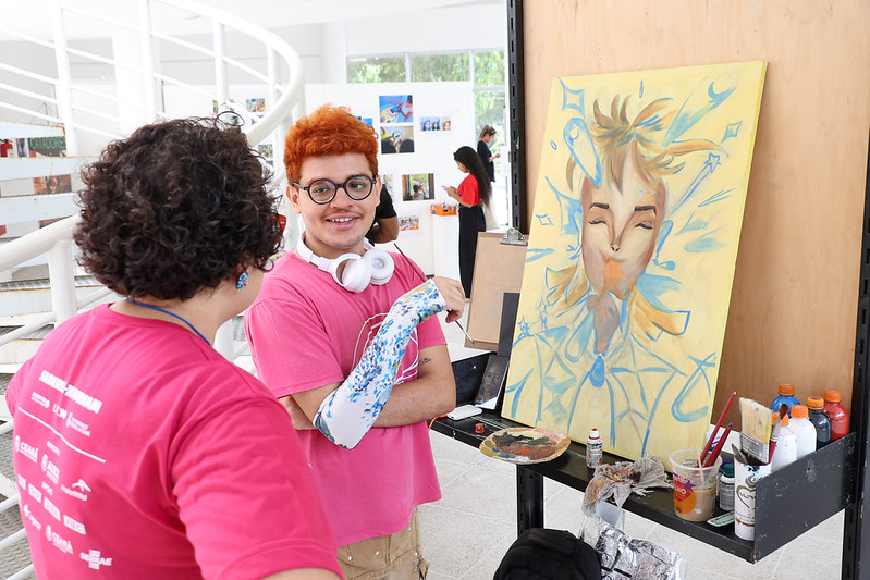As inscrições da XI Mostra ICA são abertas à toda comunidade acadêmica da UFC (Foto: Viktor Braga/Secom-UFC) Imagem: Na imagem, duas pessoas estão em um ambiente interno claro e moderno, durante a Mostra ICA 2024. Ambas vestem camisetas rosas. Em primeiro plano, uma delas — de cabelo curto e ruivo, óculos redondos e fones brancos pendurados no pescoço — está sorrindo enquanto segura pincéis. À sua frente, há uma tela apoiada em um painel, com uma pintura colorida de um rosto estilizado, em tons de amarelo e azul, cercado por formas geométricas e abstratas que lembram respingos e estrelas. Na lateral da tela, há pincéis, tintas e recipientes de apoio. Ao fundo, outras pessoas observam imagens expostas na parede branca, compondo o cenário do evento artístico coletivo.