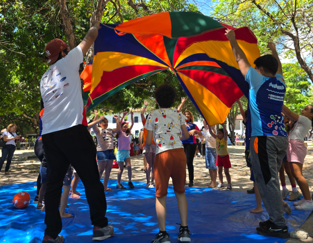 Imagem: Em um espaço aberto e ensolarado, um grupo de crianças e adultos participa de uma brincadeira coletiva com um grande paraquedas colorido. As cores vivas — laranja, verde, azul, amarelo e vermelho — formam um círculo vibrante no centro da imagem. Todos estão de pé, levantando o tecido para o alto, criando um efeito de onda. O chão está coberto por uma lona azul, e há árvores ao redor que fornecem sombra parcial. Foto: Christiane Martins/ Divulgação Casa de José Alencar)