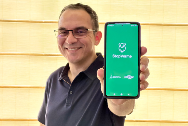 Imagem: homem de óculos e camisa polo preta posa para foto mostrando a tela de um telefone celular, onde, sobre um fundo verde, lê-se os nomes StopVerme e Embrapa e a sigla da Universidade Federal do Ceará 