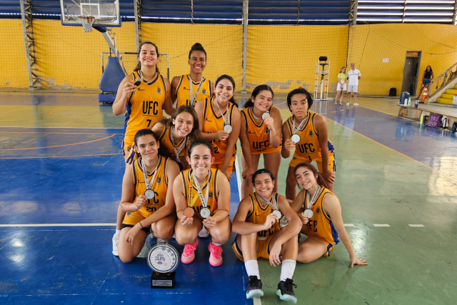 Imagem: Grupo de dez atletas do time feminino de basquete da Universidade Federal do Ceará (UFC) posa para foto em quadra esportiva coberta, com piso em tons de azul e verde e paredes amarelas ao fundo. As jogadoras, que vestem uniformes amarelos com detalhes azuis e a sigla “UFC” estampada no peito, estão sorrindo e exibem medalhas penduradas no pescoço. À frente do grupo, no chão, há um troféu preto e prateado. Ao fundo, vê-se uma tabela de basquete e parte da arquibancada.