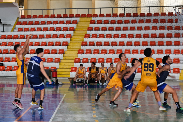 Imagem: Atletas das equipes masculina da Universidade Federal do Ceará (UFC) e da Universidade Federal da Bahia (UFBA) disputam partida de basquete em ginásio coberto. O jogador da UFC, de uniforme amarelo, arremessa a bola em direção à cesta, enquanto adversário da UFBA, de uniforme azul, tenta bloquear o lance. Outros atletas das duas equipes estão próximos da área do garrafão, posicionados para o rebote. Ao fundo, há arquibancadas com assentos vermelhos e degraus amarelos, além de jogadores reservas sentados no banco.