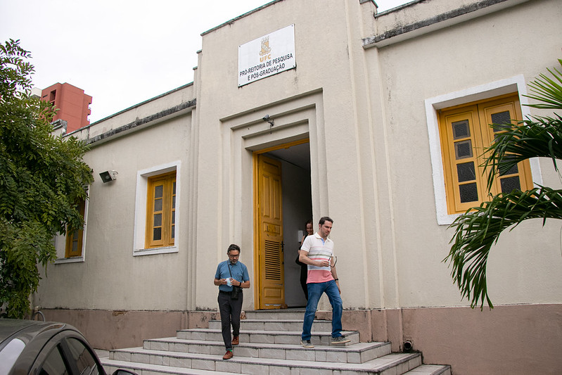 Imagem: foto da fachada do prédio da PRPPG nas cores bege com janelas amarelas ao lado de uma porta central amarela também. dois homens aparecem descendo as escadas em frente à porta central