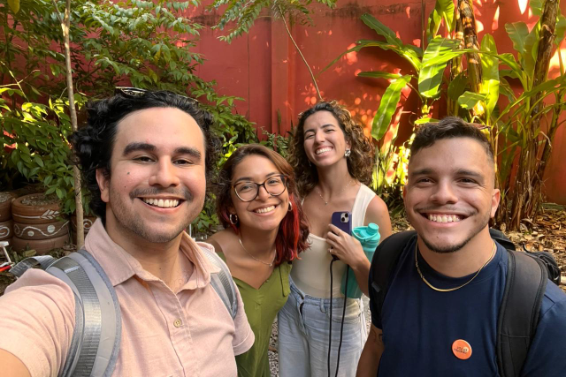 Imagem: quatro pessoas sorriem para uma selfie em um ambiente externo com vegetação e um muro vermelho ao fundo. Todos estão de mochila e parecem alegres