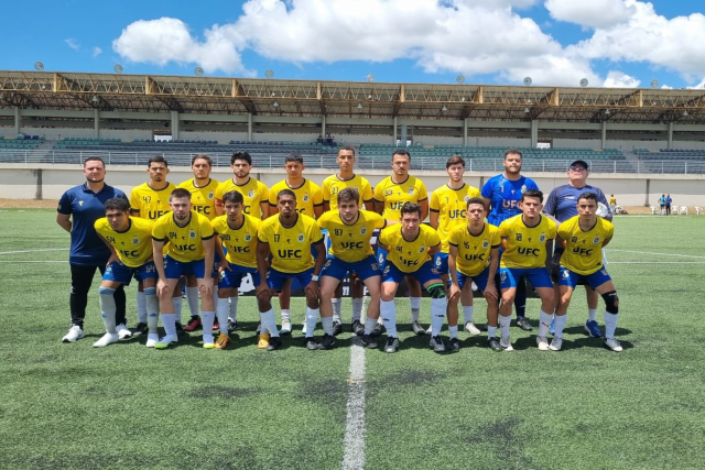 Imagem: #ParaTodosVerem: Foto do time masculino de futebol da Universidade Federal do Ceará posando em campo gramado sob céu azul com nuvens. O grupo é composto por 17 pessoas, incluindo atletas e comissão técnica. Os jogadores usam uniforme amarelo com detalhes em azul e a sigla “UFC” estampada no peito, além de shorts azuis e meiões brancos. Eles estão alinhados em duas fileiras: a da frente agachada e a de trás em pé. Ao fundo, vê-se a arquibancada de um estádio.