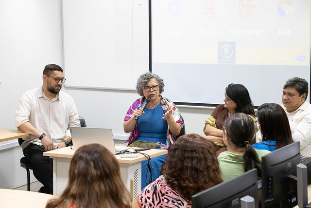 A vice-reitora da UFC, Diana Azevedo, ressaltou a importância profissional e cultural do domínio de uma segunda língua: "abre portas e gera conexões que inteligência artificial nenhuma substitui" (Foto: Lamonier Rodrigues/UFC) Imagem: encontro em uma sala de informática com diversas pessoas sentadas em círculo. Quatro participantes estão à frente conduzindo a conversa, enquanto o restante observa. Dois projetores exibem telas diferentes na parede ao fundo. O clima é de apresentação e diálogo coletivo.