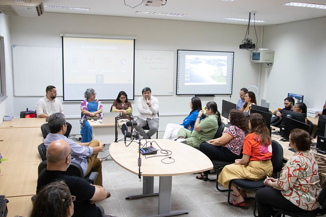 O lançamento contou com a presença de docentes e alunos dos departamentos envolvidos, além de gestores da universidade e da Funcap (Foto: Lamonier Rodrigues/UFC) Imagem: mesa-redonda em uma sala de aula. Uma pessoa ao centro fala ao microfone enquanto outras duas, sentadas ao lado, acompanham atentamente. Participantes estão sentados em semicírculo ouvindo a discussão. Há um projetor ligado ao fundo exibindo ícones coloridos relacionados a habilidades de linguagem.