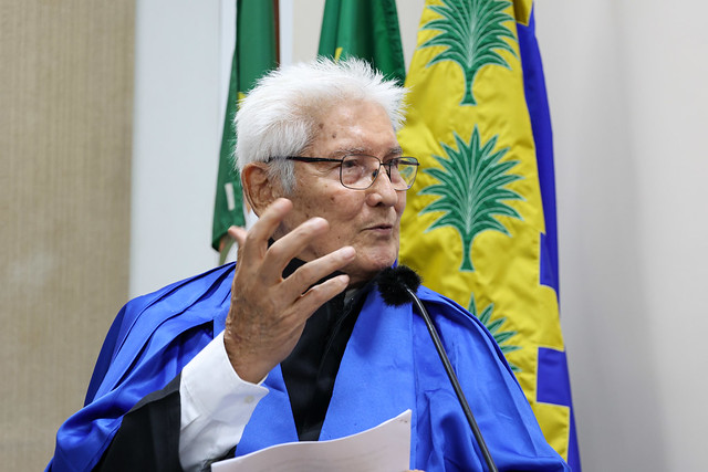Em seu discurso, o professor José Osvaldo Carioca agradeceu pelo apoio da família, lembrando das inúmeras vezes em que precisou abrir mão do convívio para dar conta das demandas acadêmicas (Foto: Viktor Braga/UFC) Imagem: Pessoa idosa com cabelo branco, usando beca azul, fala em pé diante de um microfone, segurando folhas de papel. Atrás, aparecem bandeiras oficiais presas a mastros.