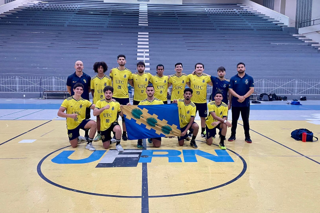 Imagem: Fotografia de um time masculino de handebol da Universidade Federal do Ceará (UFC). O grupo está posicionado no centro de uma quadra esportiva, sobre o logo pintado no chão da UERN. São treze pessoas ao todo: onze atletas e dois integrantes da comissão técnica. Na fileira da frente, cinco jogadores estão agachados segurando, ao centro, uma bandeira azul com desenhos em verde e amarelo. Na fileira de trás, seis atletas estão em pé, todos vestindo uniforme amarelo com detalhes em azul-marinho e o nome “UFC” no peito. À esquerda e à direita, dois treinadores usam camisa azul-marinho e posam ao lado do grupo. As arquibancadas ao fundo estão vazias e o ginásio é amplo e bem iluminado.