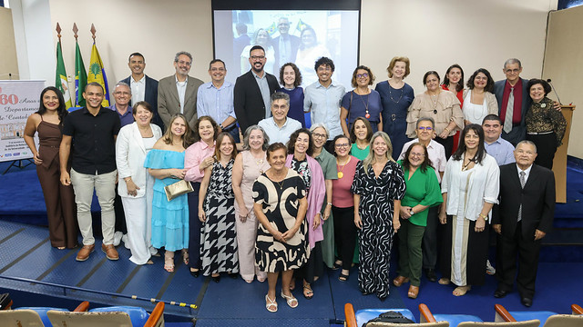 Imagem: grupo numeroso de pessoas adultas, de diferentes idades, gêneros e tons de pele, posa para uma foto coletiva no palco. Elas estão organizadas em fileiras, algumas em pé e outras à frente. A maioria veste roupas sociais ou de passeio, em cores variadas. Ao fundo, há uma mesa longa, cadeiras azuis, bandeiras oficiais posicionadas à esquerda e uma tela de projeção exibindo uma imagem do evento. O clima é formal e celebrativo, com muitas pessoas sorrindo e olhando para a câmera.