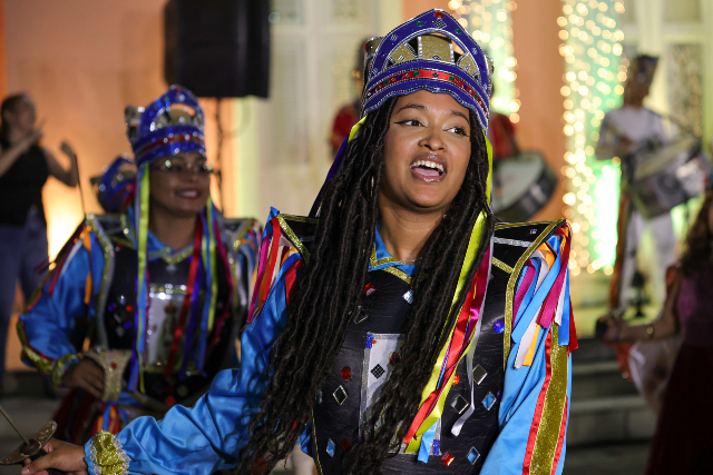 O grupo cultural Brincantes Cordão do Caroá se apresenta na terça-feira (6), a partir das 19h, nos jardins da Reitoria (Foto: Viktor Braga/ Secom-UFC) Imagem: Mulher com traje de reisado, fitas coloridas na cabeça e sorriso no rosto, em frente à Reitoria da UFC