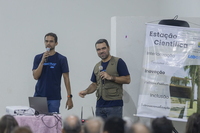 Imagem: Dois homens estão em pé em um auditório, falando ao público com microfones. À esquerda, um homem jovem veste camiseta azul escura e segura um microfone com a mão direita, ao lado de uma mesa com notebook e projetor. À direita, outro homem, também jovem, usa camiseta azul e um colete verde com bolsos, segurando um microfone e sorrindo levemente. Ao fundo, há um banner vertical com o título “Estação Científica” e palavras como “Interiorização”, “Inovação”, “Políticas Públicas”, “Inclusão” e “Internacionalização”. Em primeiro plano, aparecem as cabeças do público sentado, visto de costas.