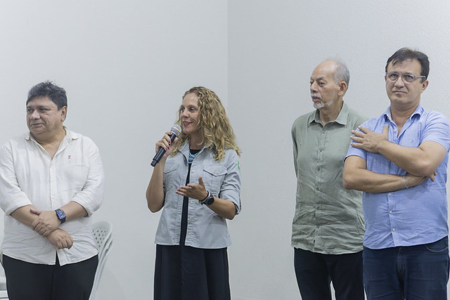 Imagem: quatro pessoas estão em pé diante de uma parede branca. Ao centro, uma mulher de cabelo cacheado e claro segura um microfone e fala, com expressão confiante. À esquerda e à direita dela, três homens observam atentamente. Todos vestem roupas casuais, em um ambiente interno bem iluminado.