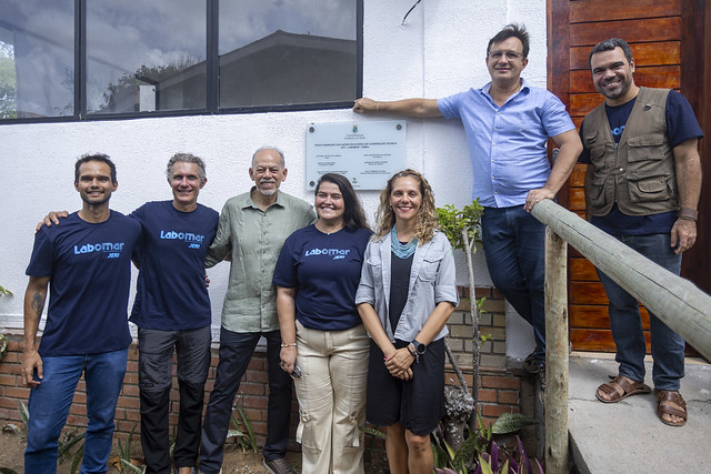 Imagem: grupo de oito pessoas posa para foto em frente a um prédio institucional. Sete pessoas estão alinhadas em pé, lado a lado, sorrindo para a câmera, e uma pessoa está apoiada em um corrimão à direita, em posição mais elevada. Algumas pessoas vestem camisetas azul-escuro com a palavra “Labomar” escrita em azul claro. No centro do grupo, há um homem mais velho de camisa verde clara. Ao fundo, na parede do prédio, vê-se uma placa comemorativa fixada. O ambiente externo inclui parede branca, detalhes em tijolo, plantas e um pequeno jardim na parte inferior da imagem.