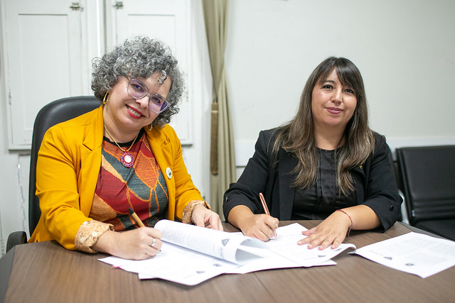 Imagem: Duas mulheres sentadas à mesa assinam documentos em uma sala institucional. Uma veste blazer amarelo e sorri; a outra usa blazer preto. Há papéis sobre a mesa.