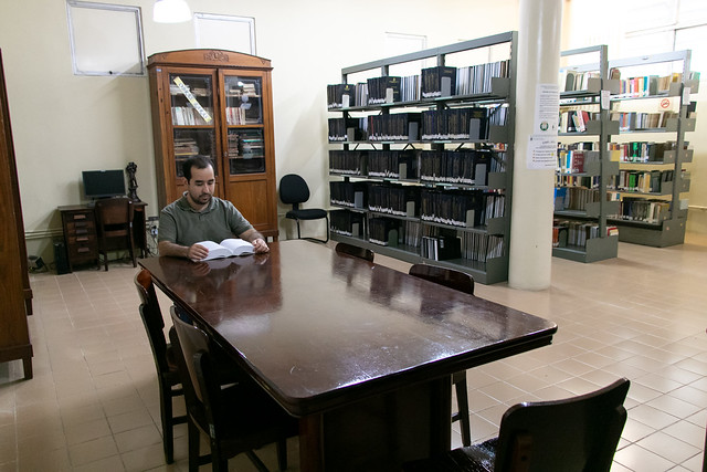 Imagem: Foto de uma biblioteca. Em primeiro plano, há uma mesa grande de madeira escura com várias cadeiras ao redor. Sentado a um dos lados da mesa, um homem adulto lê um livro aberto, com postura concentrada. Ao fundo, aparecem estantes altas repletas de livros organizados, um armário antigo de madeira com portas de vidro e uma coluna clara no centro do ambiente. O espaço é amplo, iluminado e silencioso, com piso claro e aparência de sala de estudos acadêmica.
