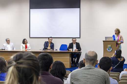 Imagem: Vista das autoridades sentadas na mesa solene, enquanto a superintendente do Complexo Hospitalar Josenília Gomes fala no púlpito. Ao fundo, uma apresentação está sendo projetada em um telão