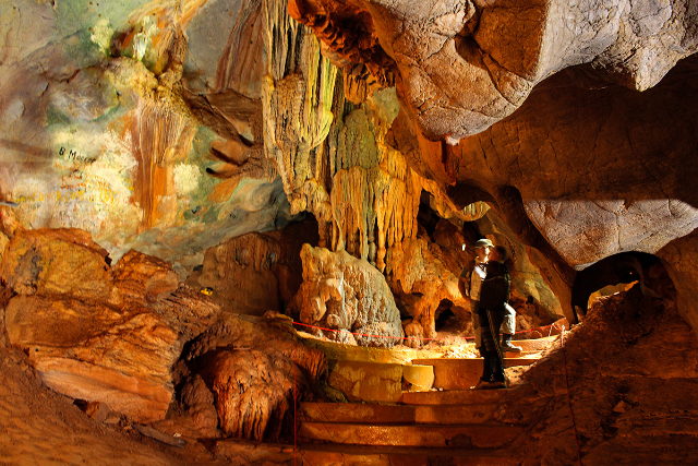 Imagem: Fotografia do interior de uma caverna com iluminação em tons quentes, predominando o laranja e o dourado. No centro e no alto da imagem, há formações rochosas com estalactites alongadas e irregulares, além de paredes com manchas esverdeadas e azuladas. Em primeiro plano, aparecem degraus de pedra que conduzem a um pequeno patamar circular protegido por uma corda de isolamento. À direita, uma pessoa usando capacete com lanterna observa as formações da caverna, em pé sobre os degraus. O ambiente é rochoso, com diferentes texturas e volumes, sugerindo profundidade e grande dimensão do espaço subterrâneo.