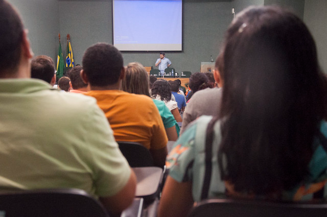 As iniciativas aprovadas contarão com apoio financeiro, incluindo bolsas e recursos de custeio (Foto: Ribamar Neto/UFC Informa) Imagem: Fotografia de um auditório visto a partir do fundo da plateia. Em primeiro plano, aparecem pessoas sentadas, vistas de costas, ocupando cadeiras enfileiradas. Ao fundo, há um palco com mesa e cadeiras, além de um telão de projeção exibindo tela azul. Um homem está em pé à frente da mesa, segurando microfone e falando ao público. À esquerda do palco, estão posicionadas bandeiras, incluindo a do Brasil. O ambiente é fechado, com paredes em tom neutro e iluminação artificial.