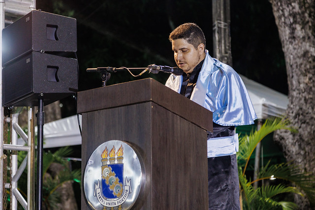 Imagem: Homem jovem, de cabelos curtos e barba leve, veste beca escura e uma capa azul brilhante. Ele está de pé atrás de um púlpito de madeira com o brasão da Universidade Federal do Ceará, falando ao microfone durante uma cerimônia noturna ao ar livre.