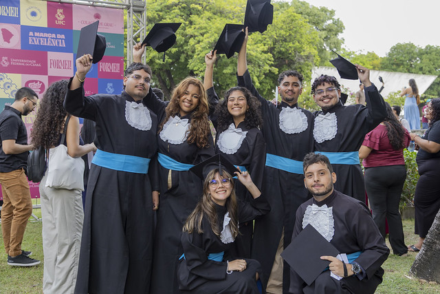 Imagem: Um grupo de sete jovens formandos posa ao ar livre durante uma cerimônia de graduação. Eles usam becas pretas com faixas azuis na cintura e detalhes brancos no peito. Cinco deles estão em pé e levantam seus capelos (chapéus de formatura) no ar, sorrindo para a foto.