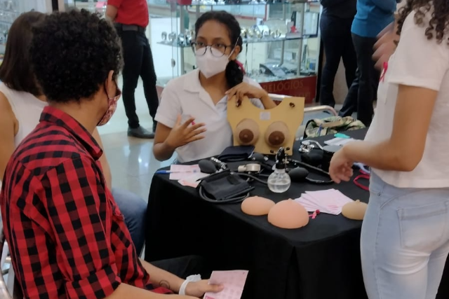 Imagem: Fotografia de uma ação educativa em ambiente interno, possivelmente um shopping ou espaço de circulação. Ao centro, uma estudante da área da saúde, usando camisa branca, óculos e máscara de proteção, conversa com duas pessoas em frente a uma pequena mesa coberta com toalha preta. Sobre a mesa, há materiais educativos sobre saúde das mamas, incluindo um modelo anatômico de tórax com seios, próteses de silicone simulando mamas e instrumentos utilizados para demonstração. À esquerda, uma pessoa sentada, usando camisa xadrez vermelha e preta e máscara, observa a explicação. À direita, outra pessoa em pé acompanha a atividade. Ao fundo, vitrines de loja com relógios e pessoas circulando pelo local.