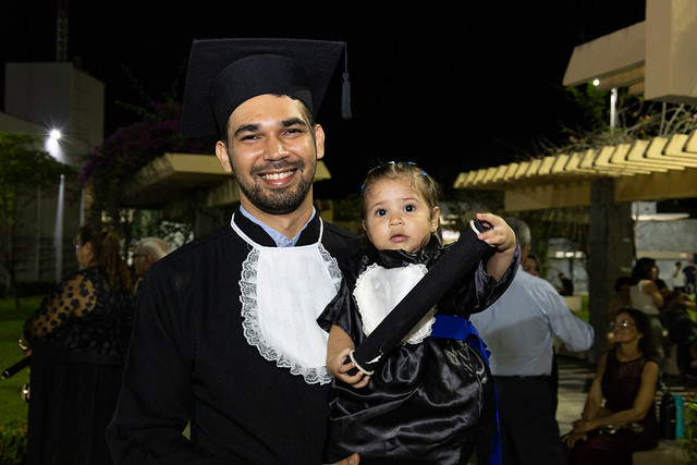 Imagem: Homem sorridente vestido com beca preta de formatura e faixa azul segura um bebê no colo. O bebê também usa uma pequena roupa preta semelhante à beca e segura um canudo de diploma. Ambos estão ao ar livre à noite.