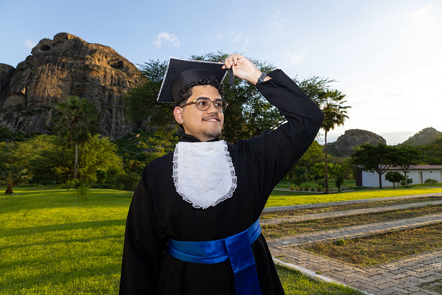 Imagem: Homem jovem usando beca preta de formatura, faixa azul e óculos está em pé em um jardim durante o dia. Ele levanta a mão para segurar ou ajustar o capelo sobre a cabeça. Ao fundo há árvores, gramado e grandes formações rochosas, com luz do sol iluminando o cenário natural.