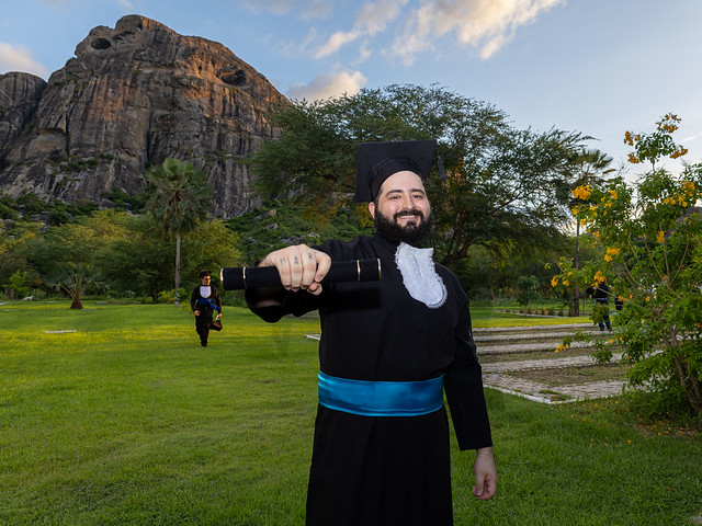 Imagem: Homem jovem, de barba e cabelos escuros, veste beca preta com faixa azul na cintura e segura um diploma enrolado com o braço estendido, sorrindo. Ele está em um gramado amplo ao ar livre.