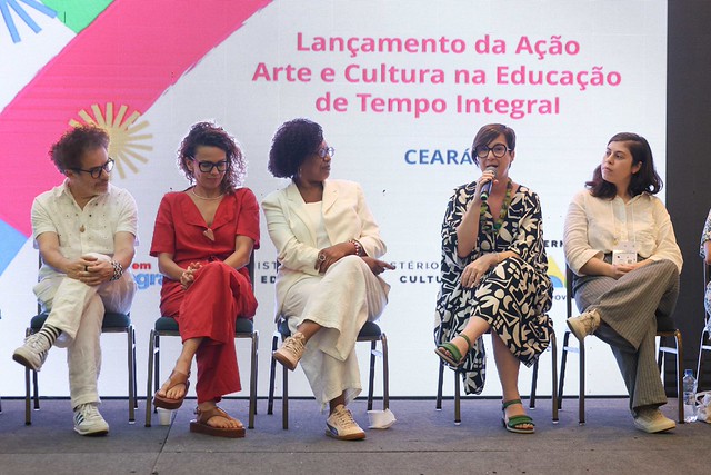 Imagem: Cinco pessoas estão sentadas em cadeiras em um palco. Da esquerda para a direita: um homem de camisa clara e tênis; uma mulher de macacão vermelho; uma mulher de terno branco; uma mulher de vestido preto e branco segurando um microfone; e uma mulher de camisa branca e calça cinza. Ao fundo, há uma tela com o texto sobre um evento de arte e cultura na educação