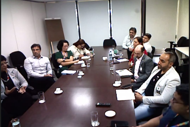 Imagem: Sala de reuniões com uma mesa retangular de madeira ao centro. Ao redor, cerca de dez pessoas adultas, de diferentes gêneros, estão sentadas, vestindo roupas formais e crachás, indicando ambiente institucional. Sobre a mesa, há copos de água, xícaras de café, pires, garrafas térmicas, celulares e blocos de anotação.  Alguns participantes estão atentos à conversa, enquanto outros fazem anotações ou observam documentos. À esquerda, uma mulher gesticula enquanto fala, sugerindo que conduz ou participa ativamente da discussão. Ao fundo, há paredes claras, cadeiras extras encostadas e um quadro ou flip chart, compondo um ambiente típico de reunião administrativa ou corporativa.