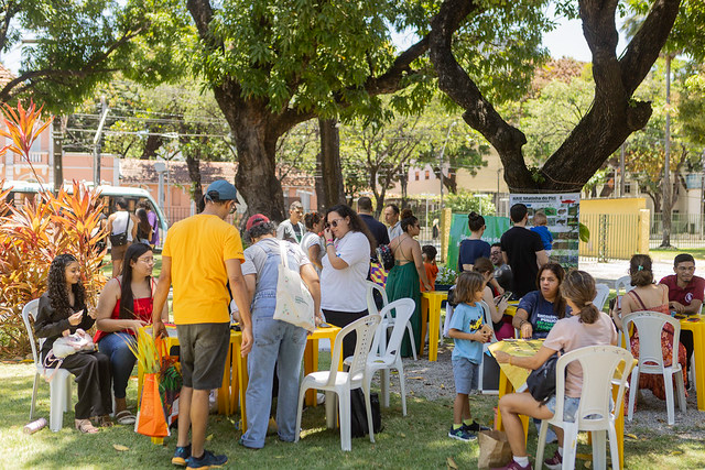 Imagem: Em área externa arborizada, várias pessoas participam de uma atividade coletiva em mesas e cadeiras de plástico distribuídas sobre a grama. Grupos conversam, escrevem e interagem em torno de mesas amarelas. Há adultos e crianças, com vestimentas casuais e coloridas. Ao fundo, vê-se um portão, placas informativas e construções do campus, sob luz natural intensa.