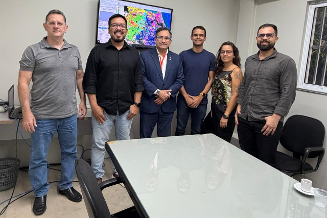 Imagem: Seis pessoas estão em pé lado a lado dentro de uma sala de escritório e atrás de uma mesa de vidro. Ao centro, um homem de terno azul e gravata sorri, cercado por colegas vestidos de forma casual, com camisas e camisetas em tons neutros. Todos estão voltados para a câmera e aparentam um clima profissional e amistoso. Ao fundo, há um monitor exibindo um mapa colorido com diferentes áreas destacadas.