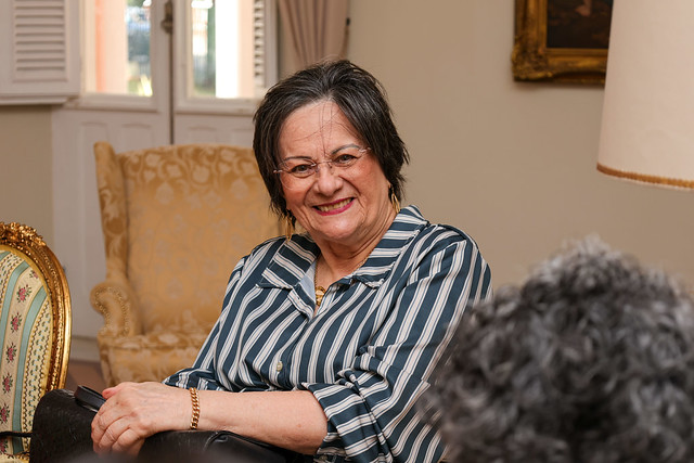Imagem: Em ambiente interno aconchegante, uma mulher de cabelos curtos grisalhos e óculos está sentada em poltrona, sorrindo para a câmera. Ela veste blusa listrada em tons de azul e branco, usa brincos e pulseira dourada. Ao fundo, há poltronas clássicas, janela com venezianas e quadro na parede. Em primeiro plano, desfocada, aparece parte da cabeça de outra pessoa.