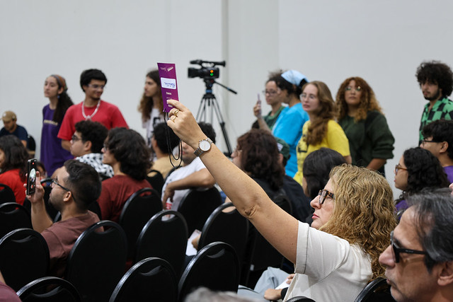 Imagem: Em auditório, uma mulher sentada levanta o braço e segura um cartão roxo durante uma votação. Ao redor, outras pessoas acompanham a atividade, algumas sentadas e outras em pé. Há cadeiras organizadas em fileiras e, ao fundo, uma câmera em tripé registra o evento. O público é diverso, com jovens e adultos em vestimentas casuais.
