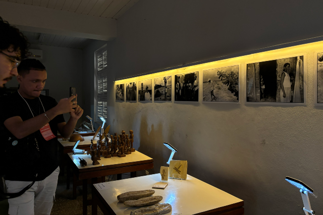 Imagem: Fotografia em ambiente interno de exposição. À esquerda, dois homens observam e fotografam as obras; um deles segura um celular na altura do rosto. Ao longo da parede à direita, há uma sequência de fotografias em preto e branco dispostas em linha, iluminadas por uma faixa de luz amarela. Abaixo das imagens, mesas expositivas apresentam objetos, como pequenas esculturas de madeira, peças alongadas que lembram fósseis ou artefatos e suportes com iluminação direcionada. O espaço é fechado, com paredes claras e iluminação suave, criando um clima de galeria.