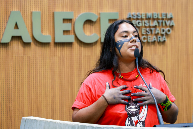 Imagem: Uma jovem indígena, com longos cabelos escuros, usa pintura tradicional no rosto e nas mãos enquanto fala ao microfone na tribuna. Ela veste camiseta vermelha e colares artesanais, com as mãos apoiadas sobre o peito em gesto expressivo